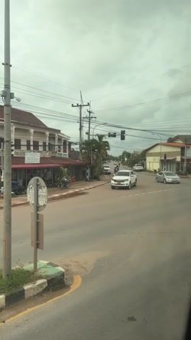 A white pickup truck turns right at a busy intersection. The sky is overcast. A traffic light is visible. Cars and motorcycles are also present on the road. Buildings line the street on either side.