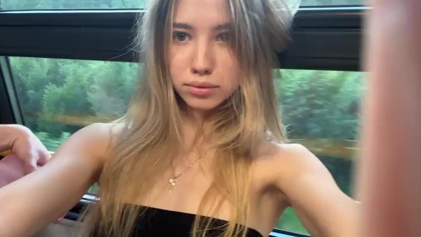 The video shows a young woman with long blonde hair, wearing a black strapless top and a necklace, sitting in what appears to be a train or bus, with a window behind her showing a blurred green, tree-filled landscape. Her right arm is bent, and a hand is visible holding a pink object. The camera, held by the person recording, is positioned in front of her face, capturing a close-up view. Her expression is neutral, and she is looking directly at the camera. The lighting is natural, coming from the window.