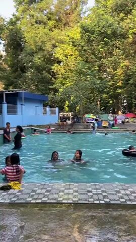 The video shows an outdoor swimming pool with several people, mostly children and young adults, swimming and playing in the water. A few adults are standing or sitting around the edge of the pool, supervising. The pool area is surrounded by lush green trees, and there are some structures visible in the background, including a blue building and several colorful umbrellas. The lighting suggests it is daytime, likely late afternoon, with sunlight filtering through the trees. Some people are using inflatable rings in the pool. One person is sitting on the edge of the pool, and another is reaching out of the water.