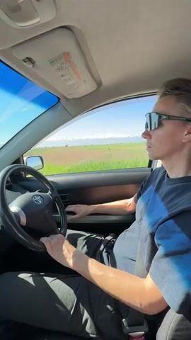 The video shows a man driving a car during the day. He is wearing sunglasses and a dark t-shirt and appears to be looking out the front windshield. The car is a Toyota, judging by the logo on the steering wheel. Outside the window, there are fields of green crops in the foreground and snow-capped mountains in the distance under a clear blue sky. The window has a long crack. The camera appears to be mounted inside the car on the passenger side. The overall scene is of a scenic drive.