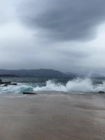 The video captures a wide shot of a turbulent sea on an overcast day in Krimovica, Montenegro. The dark, choppy ocean water, tinged with teal, crashes against a rocky shoreline and a concrete pier in the foreground. White foam and spray are visible as waves impact the land. In the middle ground, dark, craggy rocks are partially submerged by the waves. The background features a range of mountains, their peaks obscured by the low-hanging, gray clouds, suggesting stormy weather. The overall atmosphere is dramatic and wild, showcasing the raw power of nature. No people or text are visible in the frame.