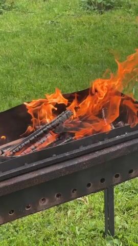 A metal brazier or grill with burning wooden logs is shown outdoors on green grass. Bright orange flames are actively consuming the wood within the brazier. The fire is substantial, with multiple tongues of flame rising from the burning logs.