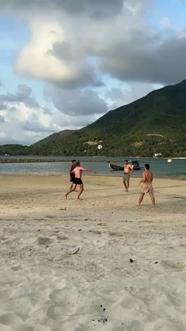 The video shows four shirtless men playing soccer on a sandy beach. The man in the center of the group kicks a soccer ball into the air. Behind them is a body of water and a densely vegetated mountain. There is a boat on the water, and the sky is partly cloudy. The overall lighting suggests it is late afternoon or early morning.