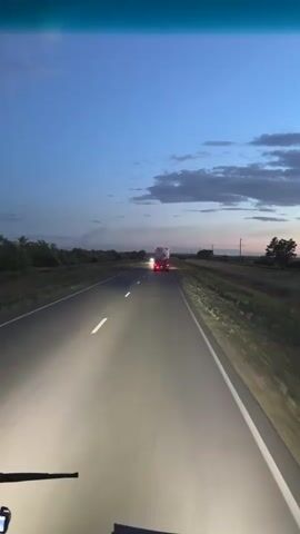 This video captures a point-of-view perspective from a vehicle driving on a highway in Baiqadam, Kazakhstan, as dusk settles. The sky is a deep blue with scattered clouds, suggesting the transition from day to night. The headlights of the vehicle are on, illuminating the asphalt road and its white lane markings.

In the distance, other vehicles are visible. Initially, a large truck with its red taillights illuminated is ahead, followed by what appears to be a car with its headlights on. Later, a car with bright headlights approaches and passes the viewer's vehicle.

The surrounding landscape consists of fields and trees on either side of the road, with distant power lines hinting at the presence of civilization. The overall atmosphere is one of travel and solitude, with the approaching darkness creating a sense of calm or perhaps mild tension. The time of day is clearly evening or twilight. No visible text is present in the video.