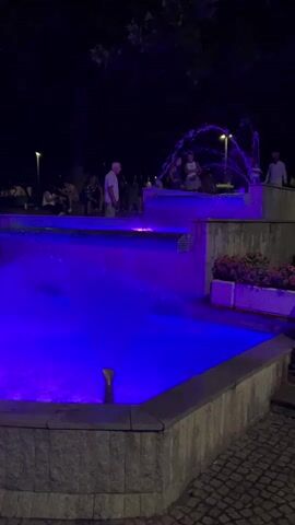 This is a vertical, night-time video of a multi-tiered fountain illuminated with vibrant blue and purple lights. Water cascades down the levels of the fountain, creating a dynamic visual effect. Several people are visible standing around the fountain, some are sitting on its edge, and a few appear to be interacting. The background is dark, suggesting an outdoor setting with trees and possibly streetlights. The overall atmosphere is festive and lively.