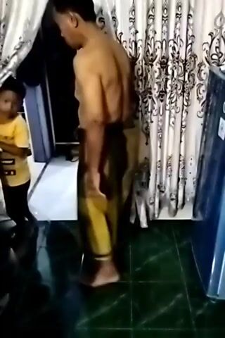 A man wearing a sarong stands shirtless in a room. He is facing away from the camera and looking towards the left. The room appears to be a living space with curtains and a refrigerator visible. Another person, partially obscured by a doorway, is also present in the scene. The floor is tiled and reflective.