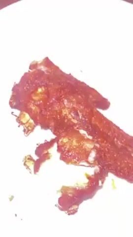 A white plate holds a partially eaten, crispy fried chicken wing. The chicken skin is a deep brown color, and a portion of the meat is exposed, revealing a pale pink hue. The camera is handheld and slightly shaky, focusing on the chicken on the plate. There are no other visible objects or people in the shot.