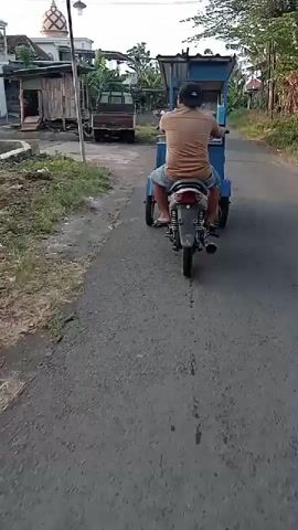 A person is riding a three-wheeled motorcycle with a blue carriage attached, down a paved road. The person is wearing a tan t-shirt and dark shorts, and a dark baseball cap. The motorcycle has a black exhaust pipe and silver trim. The blue carriage is open at the top and has a black frame. The road is lined with grass and trees. In the background, there are houses and a mosque. The sky is overcast. The camera is following the motorcycle from behind and to the right.