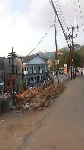 A video shows a street scene with a building that appears to be a government office or community center. A pile of rocks is on the left side of the road in the foreground. There are utility poles and wires overhead. An Indonesian flag is visible. A person rides a scooter down the street. Another person is seen running. The video appears to be filmed from a moving vehicle or by someone walking.
