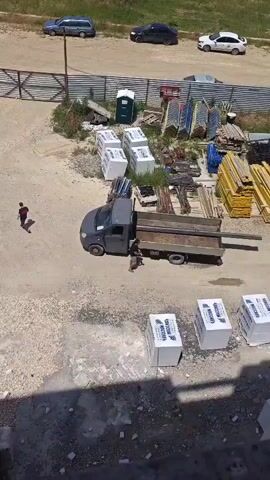 A man walks from a truck bed and then walks toward the back of a truck. He is wearing a black t-shirt, gray shorts, and sneakers. A gray pickup truck with a flatbed is parked in a dirt lot. There are stacks of construction materials, including wood and metal, in the background. A few cars are parked on a paved road in the distance.
