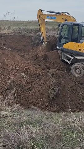 The video shows construction work in a field. A yellow excavator is digging a hole. After the excavator digs into the soil, a yellow loader tractor moves into the frame. The field is grassy and the sky is overcast. There are no people visible in the video. The excavator has "UZXCMG" written on the boom. The loader tractor is unnamed. The excavator is dumping the dirt into a large pile next to the hole.