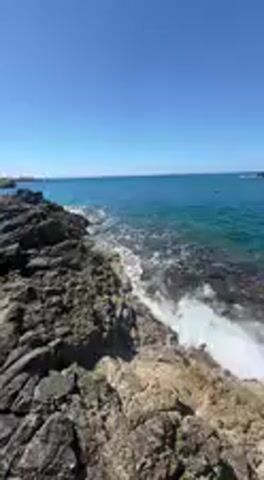This video captures a serene and picturesque rocky coastline on a bright, sunny day. The scene features dark, rugged rock formations contrasting with the vibrant turquoise waters of the ocean. Gentle white waves consistently crash and recede against the shore, creating a soothing natural rhythm. The sky overhead is a brilliant, clear blue, indicating excellent weather conditions.

At one point, the camera briefly shows a close-up of a person's bare feet and lower legs, clad in light blue trousers, standing on the rocks right at the water's edge, where the clear shallow water washes over the stones.

The perspective then returns to a wider view of the stunning seascape, highlighting the clear blue-green hues of the water and the expansive ocean stretching towards the horizon. As the video progresses, the camera pans slowly to the right, revealing more of the coastline. In the distance, a headland covered with lush green vegetation becomes visible, with a small structure, possibly a tower or building, perched on top of the cliff. The deep blue ocean extends far into the distance under the warm, bright sunlight. The overall atmosphere conveys a sense of natural beauty and tranquility, characteristic of a coastal setting like Nardò, Italy.