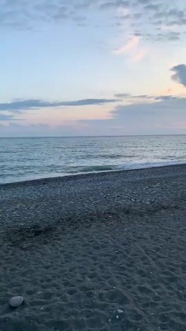 The video shows a beach at sunset. The sky is filled with clouds tinged with pink and orange hues. The sun is setting on the horizon, partially obscured by the clouds. The sea is calm with small waves lapping at the shore. The beach is made up of dark sand and pebbles. There are a few people visible in the distance walking along the beach. There are some beach chairs and what appear to be concrete blocks to protect the beach from erosion.