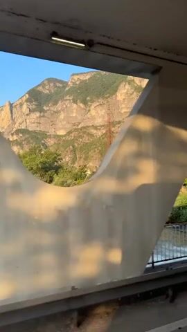The video shows a view from inside a train or vehicle moving across a bridge. Through geometric openings in the structure of the bridge, a river can be seen below, with green trees and vegetation on the banks. In the distance, a large mountain range is visible, bathed in sunlight, with a tower or pylon on its slope. The sunlight creates distinct shadows and reflections on the interior surfaces of the bridge. The railing of the bridge is visible. The overall scene depicts a scenic journey through a natural landscape.