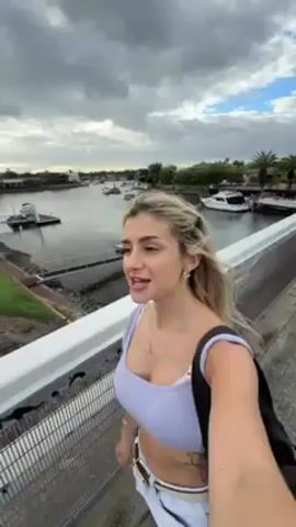A woman with blonde hair stands on a bridge. She is holding a camera to record herself and talking about having just visited the cafe where H2O was filmed and she is on her way to Cleo’s. The water behind her has boats docked. There are some houses that line the banks of the waterway. The sky is overcast. She has on a lavender top, white pants, and a gold belt. She is wearing a gold necklace and gold hoop earrings. She has a tattoo on her stomach. She has a black bag hanging over her shoulder.