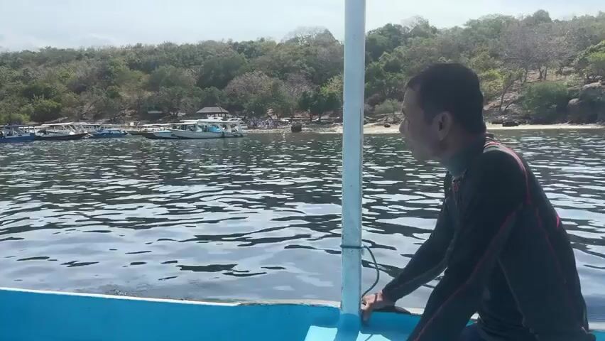 A person in a wetsuit is sitting on the edge of a boat, looking out at the water. The boat is in the ocean, and there is an island in the background with trees and some buildings. The water is calm and there are other boats anchored near the island. The sky is cloudy.