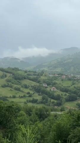 The video shows a panoramic view of a lush green mountainous landscape under a cloudy sky. The camera pans from left to right, revealing rolling hills covered in dense trees and scattered patches of grass. Several houses with light-colored roofs are visible nestled on the hillsides. As the camera continues to pan, a small town with more houses comes into view in the distance, situated within the valley. The sky is overcast, with clouds partially obscuring the peaks of the further mountains. The overall scene depicts a tranquil, rural setting.
