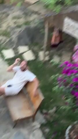 The video shows a man sitting on a wooden bench outdoors. He is wearing a white T-shirt and white shorts, and his hair is in a bun. He is holding a phone and a small orange disc. Initially, the man is facing upward with his mouth open wide, as if shouting. The camera is positioned above him, looking down.  He then glances up towards the camera and briefly speaks. The surrounding area includes stone pavers, grass, a small black table, a stone wall, and some flowers. There is also a makeshift clothes rack visible in the background.