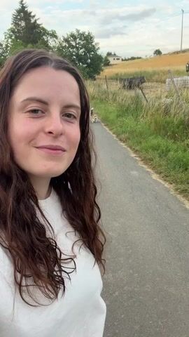 A young woman with long, dark curly hair and wearing a white t-shirt is taking a selfie on a paved path. In the background, further down the path, a small dog with brown, white, and black markings is walking away from the camera. The path is flanked by a grassy field and trees. The sky is overcast. The woman is smiling at the camera.