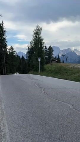 A silver car is driving towards the camera on a winding mountain road. The car's headlights are on, and it appears to be moving at a moderate speed. The road is paved with asphalt and has a white line marking the edge. Tall evergreen trees line the left side of the road, and a ski lift can be seen in the background on the right. The sky is cloudy, with hints of blue visible through the clouds. In the distance, majestic mountain peaks are visible. A circular sign with a red border and a diagonal red line through it is mounted on a pole near the road. The overall impression is of a scenic drive through a mountainous region.