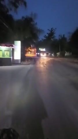A first-person perspective video captures a scene at dusk or dawn on a paved road. Trees line the left side of the road, with a brightly lit structure, possibly a guardhouse or entrance, on the far left. The road is wet, reflecting the headlights of vehicles and streetlights. In the foreground, the back of a person riding a motorcycle is visible. Further down the road, another motorcycle is seen passing by. In the distance, headlights of more vehicles can be observed, and a bus is approaching. The sky is a deep blue, indicating twilight. The overall atmosphere is that of a nighttime journey on a suburban or rural road.