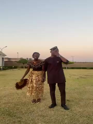 A young woman and man are standing on a grassy field, holding hands, and looking at the camera. The woman is wearing a brown and gold traditional Nigerian outfit. The man is wearing a brown traditional outfit with a matching hat. The background consists of a low wall, trees and a building. The sky is a pale pink and blue indicating twilight. The couple seems happy, and they are likely posing for a photo or video. The lighting is soft and even, typical of early evening.