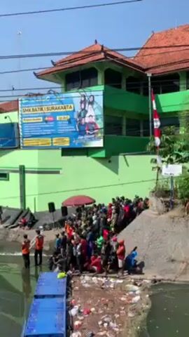 A crowd of people is gathered on the bank of a narrow waterway, in front of the SMK Batik 1 Surakarta building. The building is painted green and has a red-tiled roof. There is a large sign advertising the school on the building's exterior. An Indonesian flag flies nearby. Several people are standing in the water. There is a blue floating platform in the water, near the crowd. An umbrella is positioned in the midst of the crowd. The sky is clear and blue.