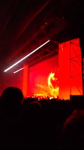 The video shows a concert viewed from the audience, with a stage illuminated by red lights. A large, spherical object is visible on the stage, as well as lights moving around.  The ceiling has metal structures and lights. The crowd is dark and only silhouettes are visible.