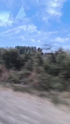 The video shows a blurry view from a moving vehicle, likely a car or bus, looking out the side window. The scenery consists of green trees and bushes, with a dirt or gravel road in the foreground. The sky is blue with some white clouds. Power lines are visible against the sky. The motion blur indicates the vehicle is traveling at a moderate speed. There are no people or distinct objects visible in detail due to the blur and the perspective.