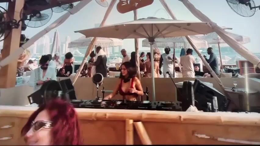 The video shows a woman DJing at an outdoor beach club. She is wearing a red top and white bottoms and standing behind a DJ setup with Pioneer equipment. There are many people in the background, some in swimwear, and the club has a tropical, beachy vibe with white umbrellas and bamboo structures. Fans are positioned around the DJ booth. The scene suggests a vibrant and lively beach party.