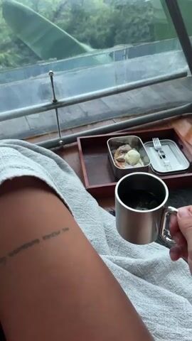 The video shows a person inside a transparent dome structure, possibly a glamping tent, with a view of a lush green landscape. The person is sitting under a gray blanket, holding a metal mug filled with a dark liquid, likely tea or coffee. A small wooden tray rests on the blanket, containing a metal container with what appears to be a pastry topped with a scoop of white ice cream, and a set of cutlery in its own metal container. Outside the dome, there is a dense tropical forest or rice field, visible through the triangular glass panels of the structure. The sky is overcast. The focus is on the scene inside the dome and the view beyond. A visible tattoo is on the person's arm.
