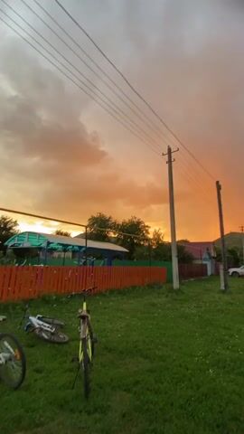 The video pans across a rural landscape during what appears to be sunset or a dramatic weather event. In the foreground, a grassy lawn is visible with a bicycle lying on its side. A brightly colored fence, predominantly orange and green, separates the lawn from a dirt road. Power lines are strung across the scene, supported by utility poles. In the background, houses with visible roofs and some vegetation are scattered throughout the area, with rolling hills in the distance. The sky is a prominent feature, displaying a vibrant orange hue on the left, transitioning to a lighter grey with clouds on the right. A faint rainbow arcs across the sky in the later frames. A white SUV is parked on the side of the road in the middle distance. A pedestrian crossing sign is also visible. The overall impression is one of a tranquil, possibly stormy, evening in a rural setting.