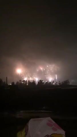The video opens with a nighttime scene of a fireworks display. The fireworks are exploding in a series of bright bursts across the sky, illuminating the clouds and smoke. In the foreground, a dark silhouette of trees and possibly buildings is visible. At the very bottom of the frame, a portion of a pink umbrella with yellow designs is visible, suggesting it is raining or has recently rained, and the camera is positioned behind it. The display appears to be taking place over a body of water or a flat, open area. The sound of fireworks is audible throughout the clip.