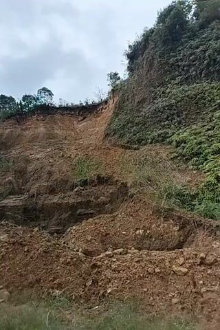 A drone flies forward, filming a large landslide on a steep, forested hillside. The landslide has deposited a significant amount of earth and debris onto the side of a road. The sky is overcast.
