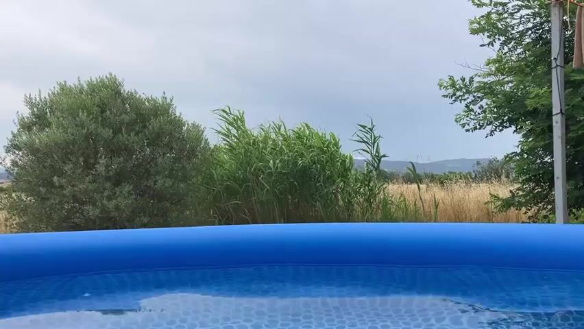 A low-angle, static shot frames the foreground of a blue inflatable swimming pool, its water rippling slightly and reflecting a hexagonal pattern on its bottom. The midground is dominated by lush green foliage, including an olive tree on the left and tall reeds in the center. Beyond the vegetation, a field of dry grass stretches into the distance, bordered by more trees and a hazy, overcast sky. The scene conveys a sense of a serene outdoor setting, possibly in a rural area.