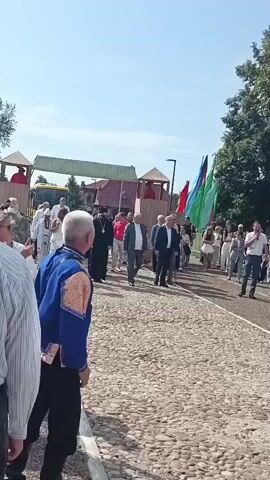 The video shows an outdoor event with a crowd of people. In the foreground, a man in a blue and gold traditional outfit is seen from behind. In the middle ground, a procession is taking place, led by a person in a black clerical robe, followed by several men in suits. Behind them, people are walking and holding colorful flags. To the right, a group of young women in traditional blue and orange dresses with red headbands are standing and facing away from the camera. Behind them, a man is speaking into a microphone. The background consists of trees, a wooden building with a thatched roof, and a clear blue sky. The ground is paved with cobblestones.