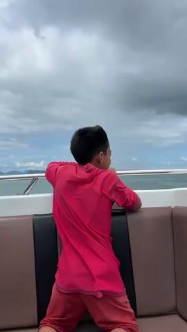 A boy with black hair, wearing a bright pink t-shirt and red pants, stands on a boat, his back to the camera. He is looking out at the ocean and a group of islands in the distance under a cloudy sky. The boat has brown and black cushioned seating visible in the foreground.