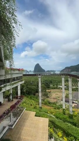 The video pans across a wide landscape. In the foreground, a building with white railings and abundant plants, including hanging vines and potted flowers, is visible on the left. Below, a section of a wooden deck is seen. A large, elevated glass walkway supported by white pillars extends across the middle ground of the frame, leading towards a body of water. Beyond the walkway, a lush green valley with agricultural fields is apparent. In the background, several islands with distinctive mountain shapes rise from the sea under a sky filled with fluffy white clouds. A person is briefly visible walking on the far right end of the glass walkway.