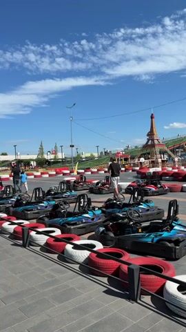 The video shows a go-kart track on a sunny day with a clear blue sky and scattered clouds. In the foreground, several go-karts are lined up, appearing to be ready for use. They are mostly black and blue with red and white tires lining the track. Two men are visible in the middle ground, one walking and the other standing near the go-karts. In the background, there is a small replica of the Eiffel Tower, and what appears to be an amusement park or entertainment venue with various structures and colorful elements. The track itself is defined by red and white barriers. The overall scene suggests an outdoor recreational area.