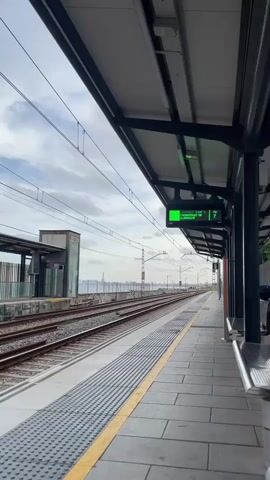 A static outdoor shot captures a train platform under a cloudy sky. The platform features two sets of train tracks, a yellow tactile paving strip, and a ribbed gray surface. Overhead power lines crisscross the frame, supported by utility poles. A digital sign above the platform displays green text reading "R1" and "09:59," along with other illegible text in Spanish or Catalan, which changes to blue text that is also illegible. In the distance, a body of water separates the tracks from a cityscape, with a prominent tall building visible. The sound of a public announcement in Spanish or Catalan plays in the background.