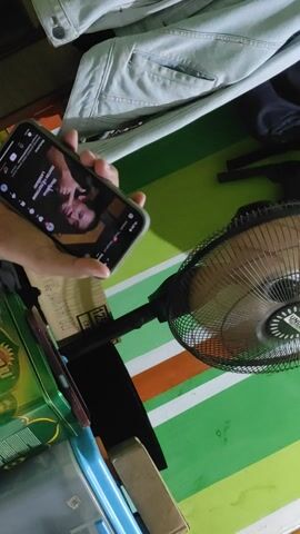 Here is a detailed description of the video:

The video shows a person holding a smartphone, viewing a video on the TikTok app. The screen displays a man speaking directly to the camera, with the caption "SIAPA YANG PALING MANLY?".  In the background, there's a wall painted with green, white, and orange stripes. A black standing fan is positioned near the wall. In the foreground, there is a blue denim jacket hanging from a chair. A green tin box and a blue storage container are also visible on the lower right side of the frame.
