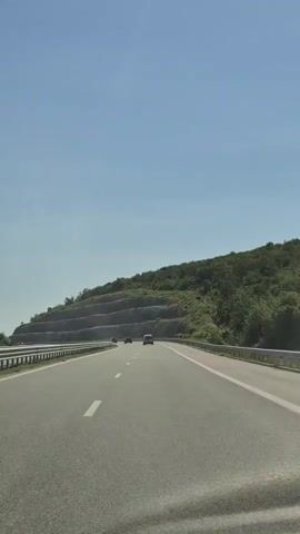 The video shows a first-person perspective of driving on a highway with a clear blue sky overhead. The road curves to the right, with a lush green hillside on the left. The hillside has been terraced with man-made retaining walls. Guardrails run along the side of the road. Several cars are visible on the highway ahead. The sun is shining, indicating it is daytime.