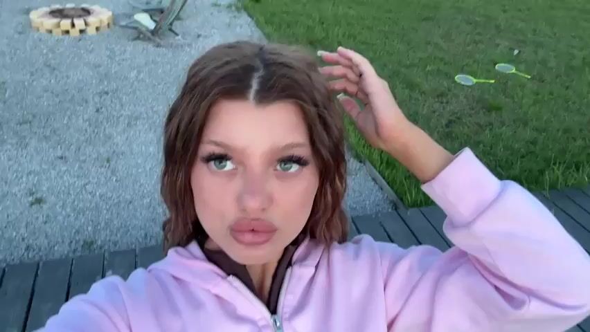 A woman is taking a selfie outdoors. She's wearing a light pink hoodie and has long, wavy brown hair. She has blue eyes and large lips. She is standing on a wooden deck next to a gravel area and a patch of green grass. In the background, there is a fire pit and two badminton rackets on the grass. She is looking at the camera, adjusting her hair, and then extending her arms to the sides.