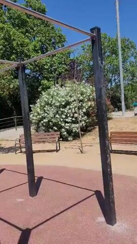 The video shows a man using a set of parallel bars at an outdoor fitness park. The park has a red rubberized floor, and the bars are made of black metal posts with silver horizontal bars. There are also some additional metal ladders on the sides of the bars. There are benches, trees, and bushes in the background. The man is wearing a white t-shirt with a logo on the back and dark pants. He jumps up to grip the bars and then uses them to support his body.