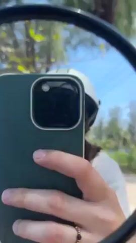 A person wearing a white helmet and sunglasses is seen taking a selfie in a motorcycle mirror. The person is holding a dark green phone with three camera lenses. The background shows a rocky shoreline with a calm turquoise sea and a clear blue sky with sparse clouds. Some trees are visible on the left of the frame. The person is wearing a beaded ring on their finger.