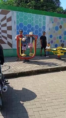 The video shows an outdoor public space with exercise equipment. In the foreground, a man assists two young children on a red and yellow swinging stepper machine. To their right, a man stands with a younger child near a yellow seesaw-like piece of equipment. Further to the right, another person uses an elliptical machine. The area is paved with light-colored brick. A wall painted with various geometric patterns and a green border forms the background. Trees are visible above the wall. A sign reading "RUANG PUBLIK OLAHRAGA" is also present on the wall. The sky is clear and blue. The scene appears to be a sunny day at a public exercise park.