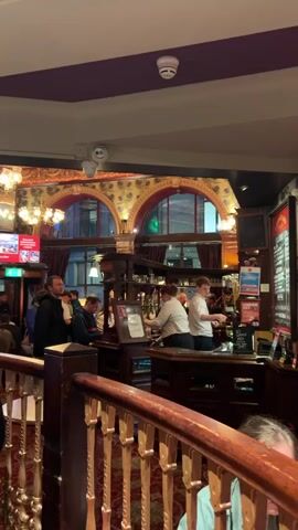 The video shows the interior of a busy pub. In the foreground, a wooden railing with gold accents separates the viewer from the rest of the pub. Behind the railing, people are standing and queuing at a bar. A bartender, wearing a grey shirt, is serving customers. Another bartender, wearing a white shirt, is also behind the bar. There are several beer taps and a menu board visible. The pub has ornate decorations, including patterned wallpaper and arched windows. The lighting is warm and inviting. People are casually dressed. The overall atmosphere appears lively and social.
