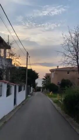 A long, narrow, paved road, surrounded by houses and vegetation on either side, leads directly to the ocean in the distance. The road slopes slightly downwards as it approaches the sea. The sky is filled with clouds, tinged with orange and pink hues, suggesting either sunrise or sunset. Power lines stretch across the sky above the road.