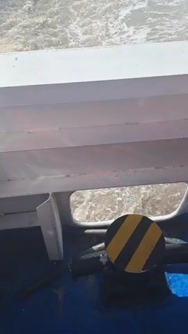 The video shows a turbulent body of water, likely the wake of a boat, viewed from the perspective of someone on the vessel. The water is a murky, brownish-gray color and is churning with white foam and waves. The perspective is looking down and slightly backward from the boat. Part of the white exterior of the boat is visible in the foreground, along with a portion of a window or opening. A black circular object with yellow diagonal stripes is positioned in the lower right corner of the frame, appearing to be a caution sign or marker. The overall impression is of movement through rough water.
