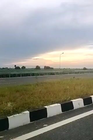 The video shows a roadside view of a highway. The sky is overcast with hints of sunrise or sunset. A light pole stands by the side of the road, and green vegetation lines the median and shoulder. The road itself appears to be a multi-lane highway with white markings. A black and white striped barrier is visible along the edge of the road in the foreground. There are no people or vehicles visible in the frame.